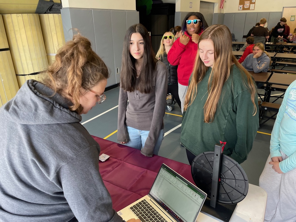 A staff member enters information on a laptop at a table while two students stand in front of a spinning prize wheel, waiting to participate. Other students are gathered in the background of the gym.