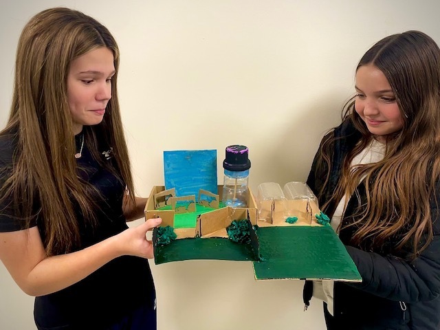 Two middle school students hold a cardboard model of their future city, featuring green-painted terrain, small structures, and a plastic bottle tower.