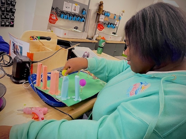 A student in a turquoise sweatshirt adds pieces to a colorful model city made of paper columns and balloons.