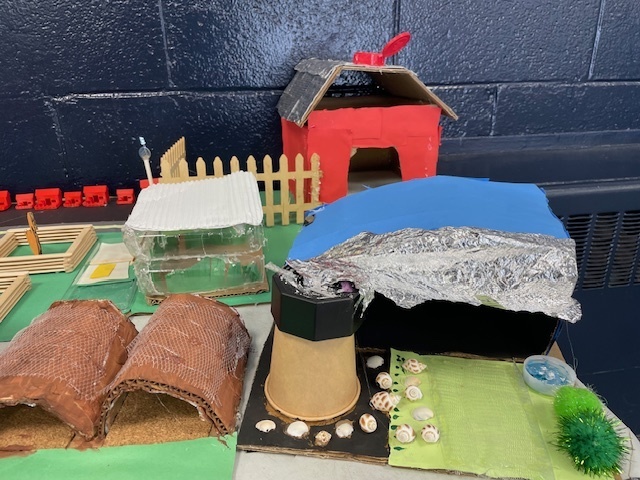 Close-up of several model structures including a red barn, clear greenhouse, brown domes, and a blue-roofed building with foil terrain.