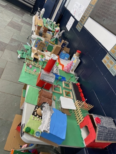 A long display table filled with many student-built future city models featuring barns, gardens, green spaces, and small structures.