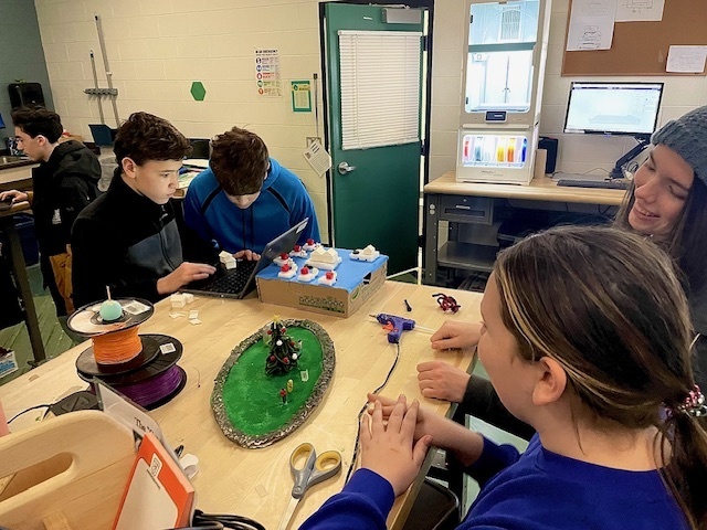 Groups of students work on their future city models in a classroom, using cardboard, paint, and craft materials spread across the tables.