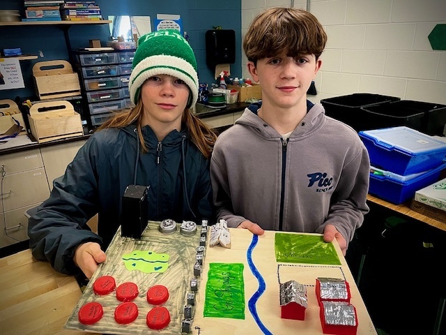 Two students proudly display a model city with roads, green spaces, bottle-cap buildings, and small red structures.