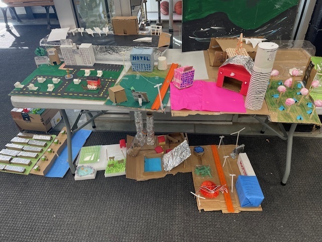 Multiple future city models displayed on tables, showing a variety of student-made structures, landscapes, and imaginative designs.