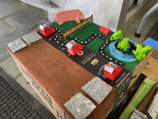 A model farm scene on a cardboard base with small red buildings, foil-covered structures, a painted road, and a pond with green model frogs.