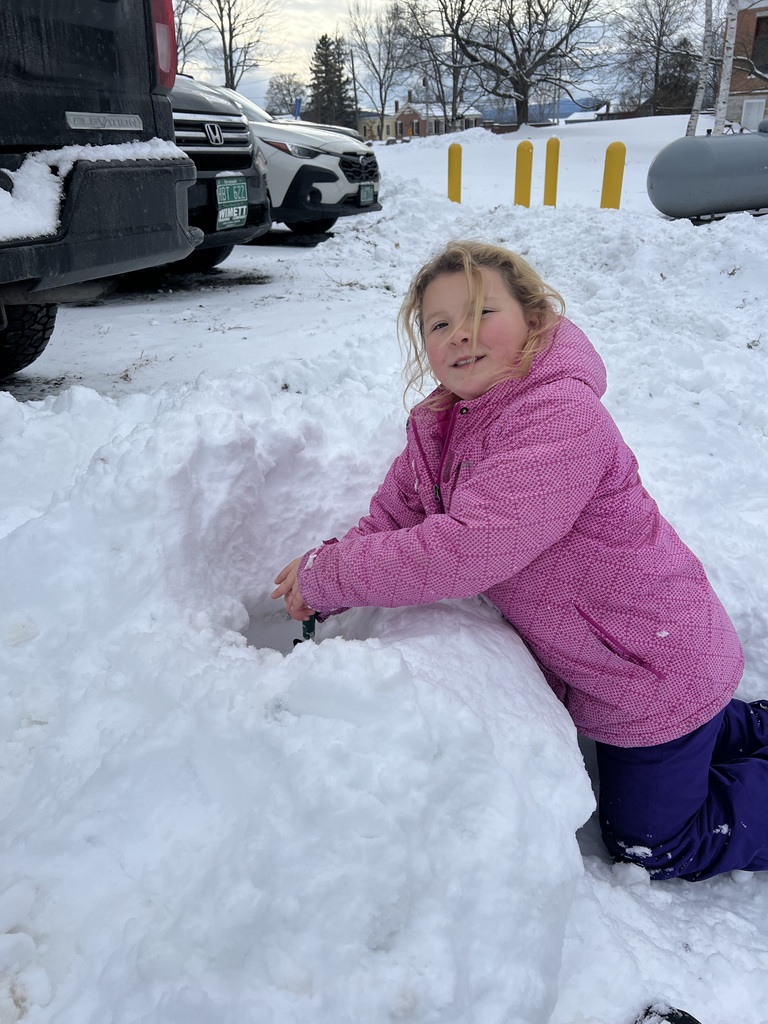 A student in a pink winter coat kneels beside a snowbank, carving into the snow with gloved hands and smiling up at the camera.