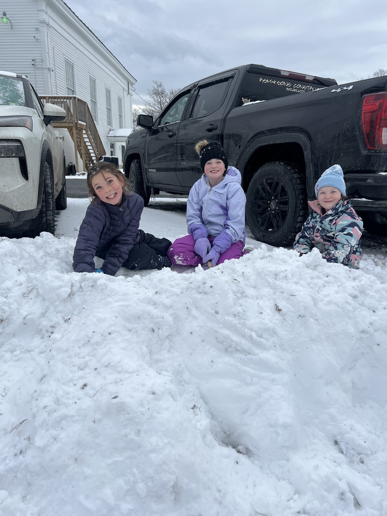Three young students sit in the snow in a parking area, bundled in winter coats and hats, smiling at the camera with trucks and a school building behind them.