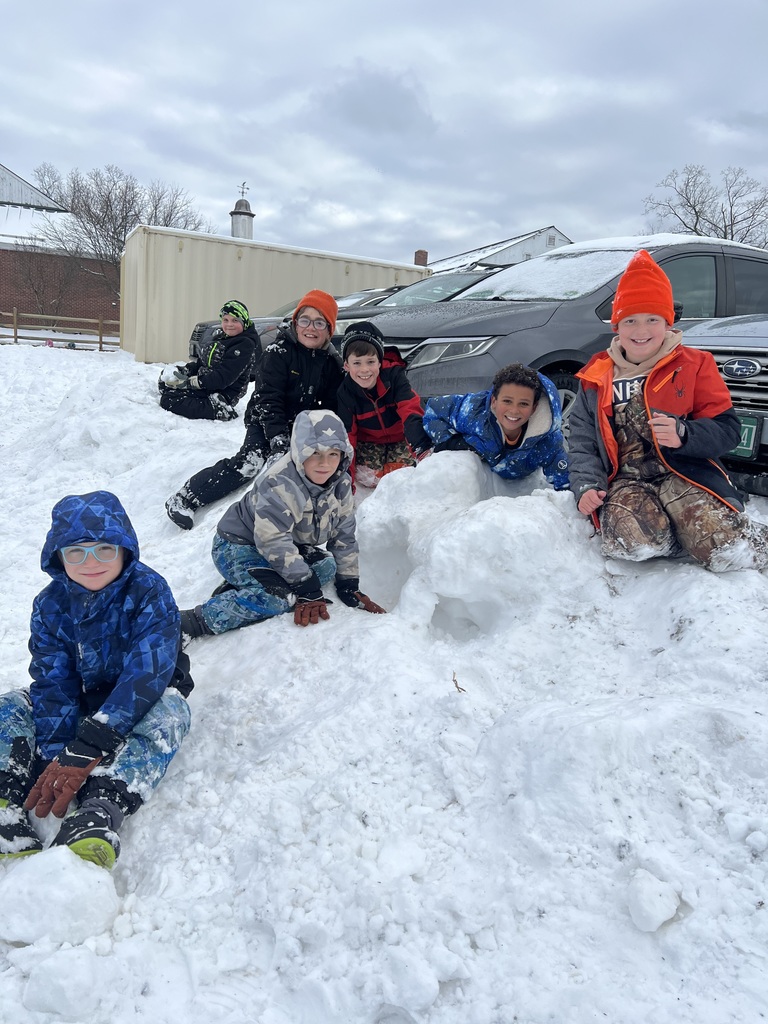 A larger group of students gather around a snow tunnel, smiling and posing proudly after building it together in the snow.