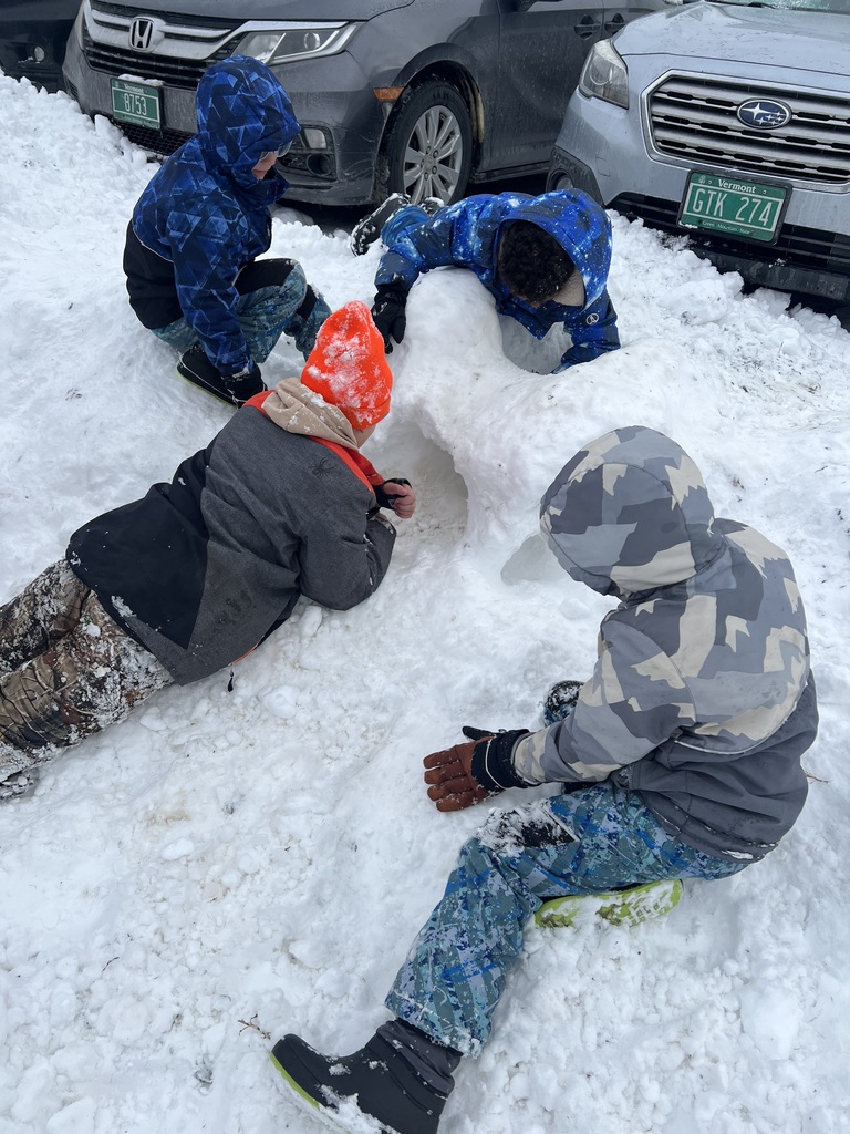 Four students in winter gear crouch and lie around a snow tunnel they are building, with parked cars in the background.
