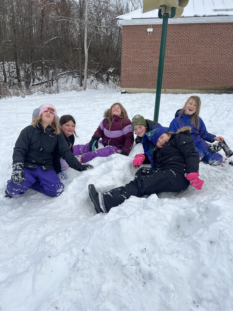 A group of elementary students sit and lie on a snowy mound outdoors, smiling and laughing together during recess near a school building.