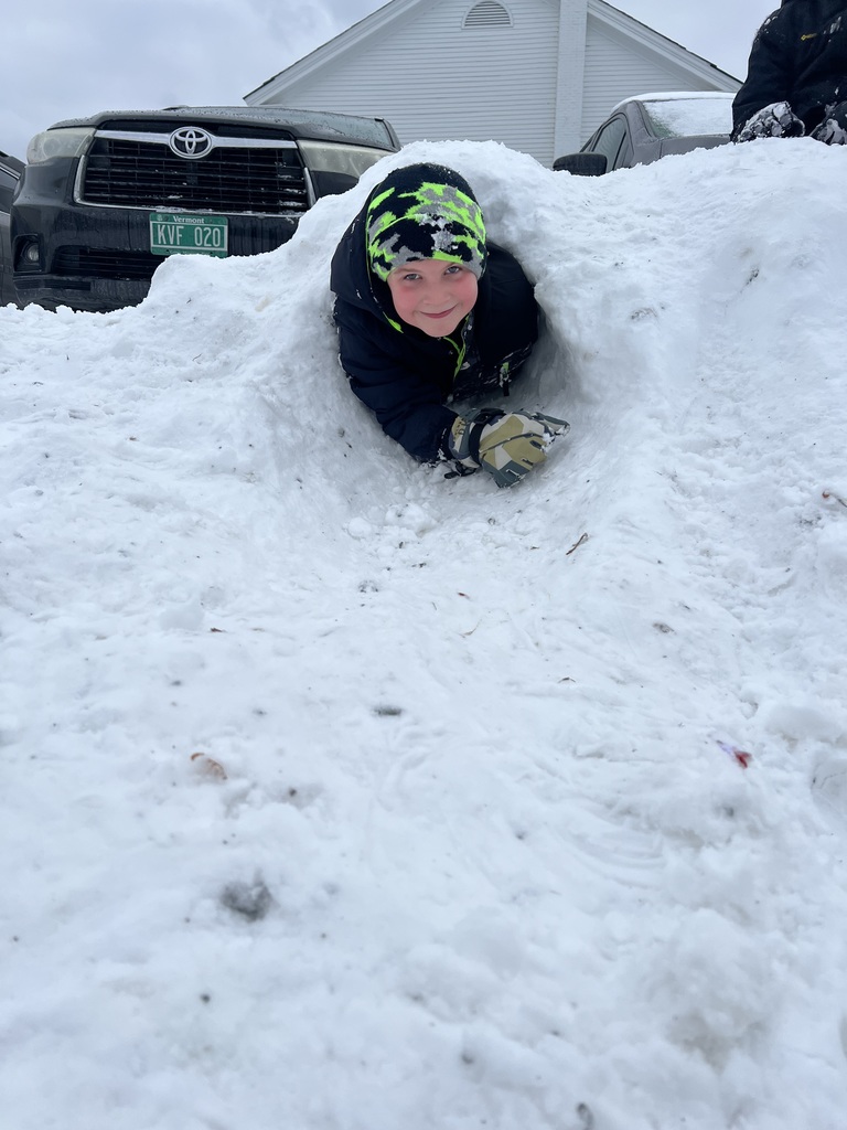 A student crawls through a snow tunnel, smiling at the camera, with parked vehicles and a snowy lot behind them.