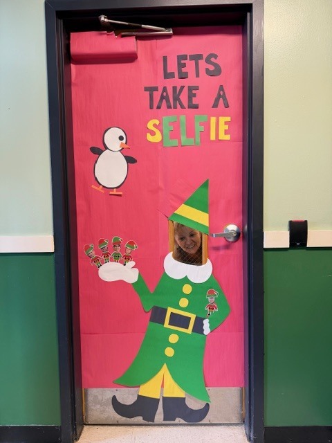 Decorated classroom door with a pink background featuring a large paper elf cutout with a face hole for selfies. The text reads “Let’s Take a Selfie,” with a small penguin graphic and festive holiday decorations.