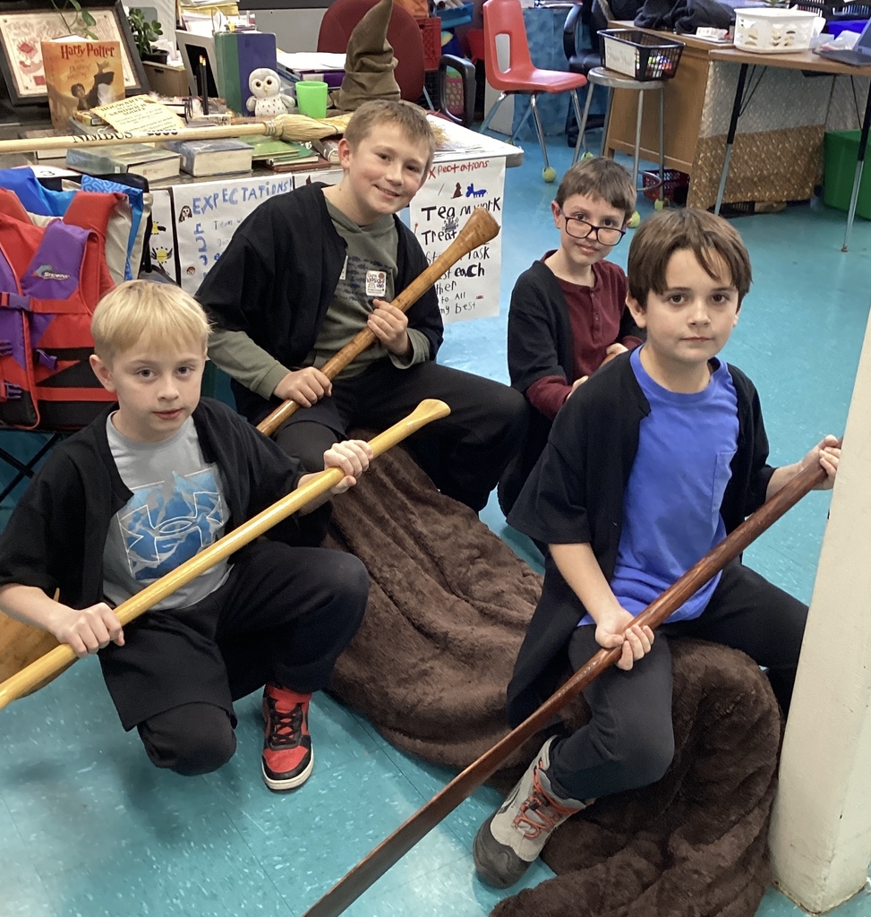 Four boys hold paddles, sitting on a brown blanket in a classroom.
