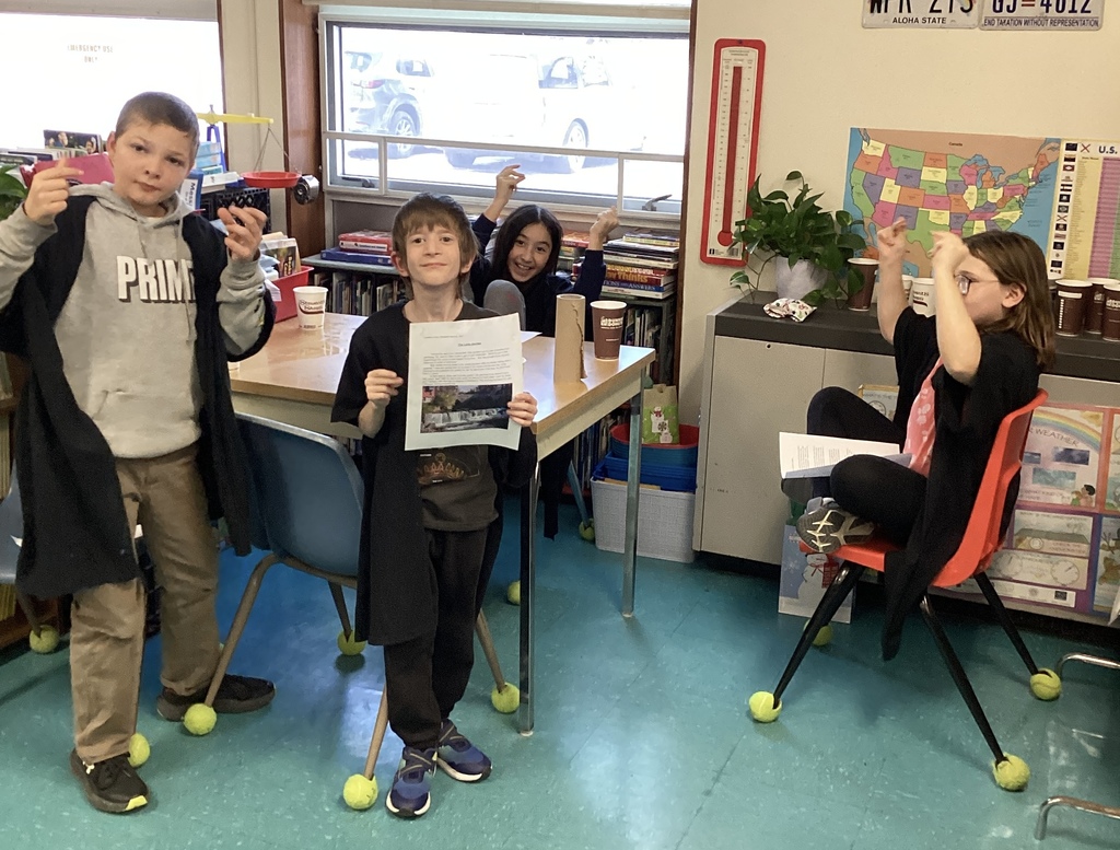 Four children in a classroom, one holding a paper and one sitting in a chair.