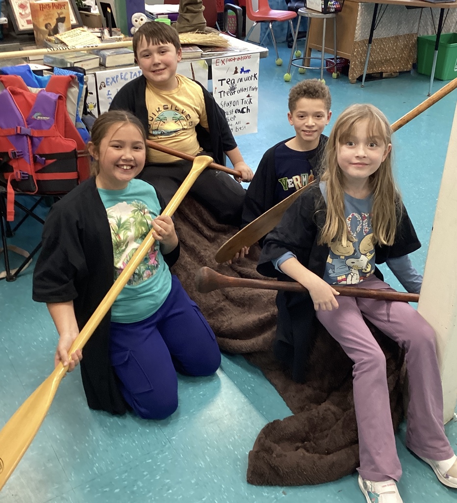 Two girls and two boys hold paddles, sitting on a blanket in a classroom.