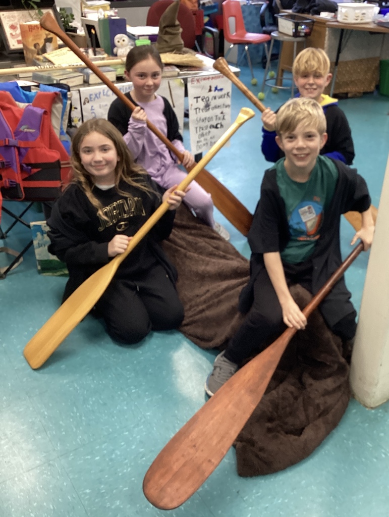 Four children hold paddles, sitting on a brown blanket in a classroom.