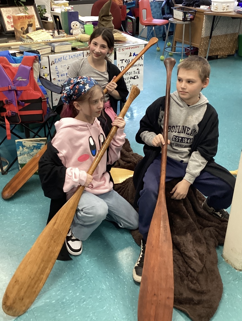 Two girls and one boy hold paddles, sitting on a blanket in a classroom.