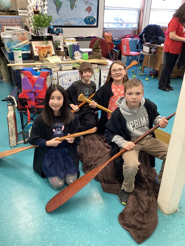 Four children hold paddles, sitting on a brown blanket in a classroom.