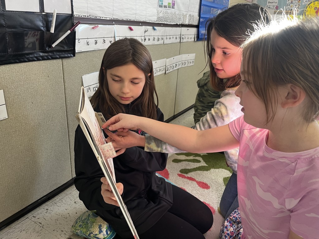 Three young girls are gathered closely together on the floor, focusing on a book. The girl in the center holds the book, and another girl points to a detail on the page, engaging in shared reading. IMG_5717.jpg: Two young girls sit on the floor of a classroom, surrounded by storage bins and books. The girl on the right, wearing overalls, holds an open book and smiles at the girl on the left, who is wearing a red outfit and looking at the book. A book titled "What About Worms?" lies on the floor nearby.