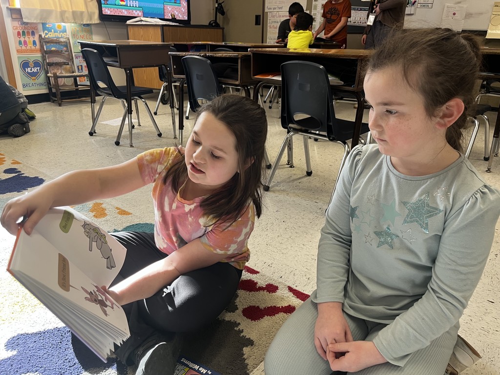 A young girl in a tie-dye shirt sits on a colorful classroom rug, pointing at a book's illustration while reading aloud to another young girl who is listening intently. Other students and desks are visible in the background.
