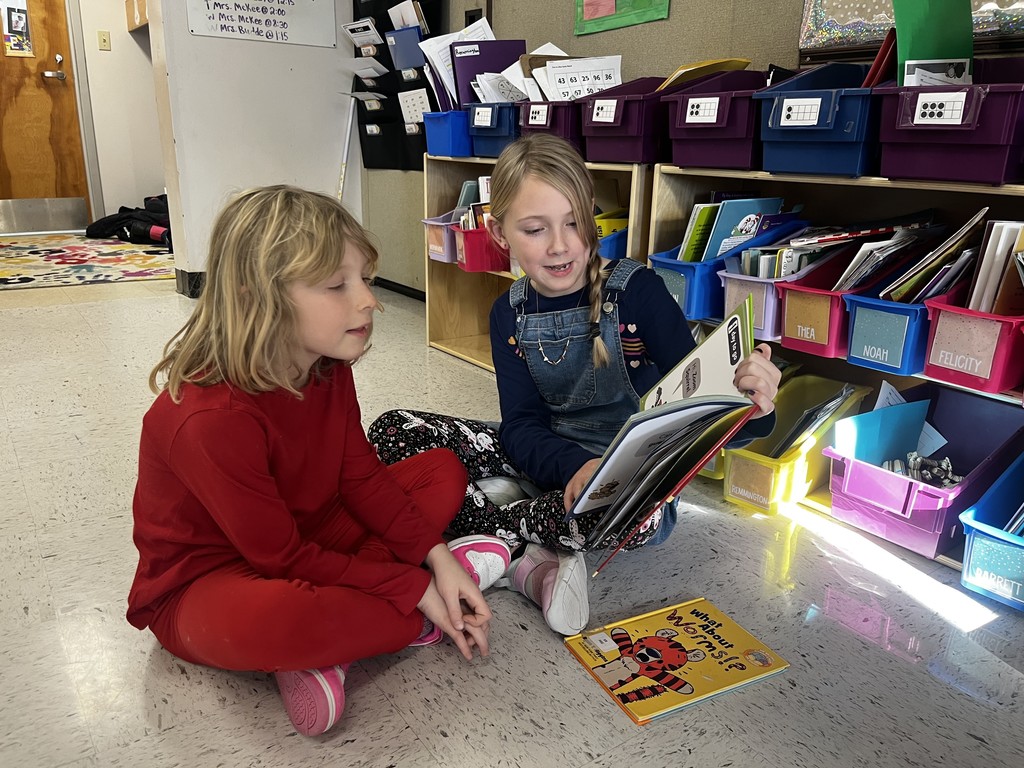 Two young girls sit on the floor of a classroom, surrounded by storage bins and books. The girl on the right, wearing overalls, holds an open book and smiles at the girl on the left, who is wearing a red outfit and looking at the book. A book titled "What About Worms?" lies on the floor nearby.