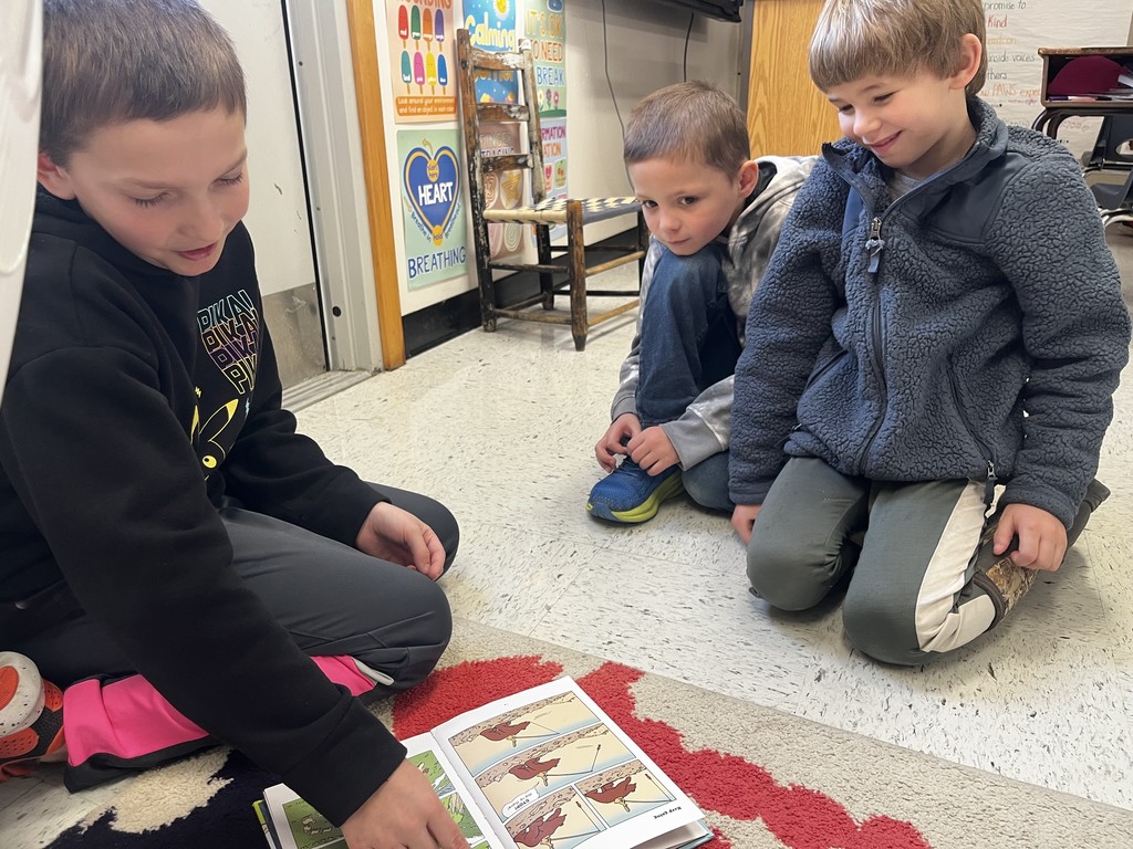 Three boys are sitting in a circle on a classroom rug, focused on a graphic novel or comic book laid flat on the floor. The boy on the left, wearing a black hoodie with pink accents, points to a panel in the book. Two other boys—one in a grey hoodie and one in a textured blue fleece—lean in with smiles, watching the story unfold. Classroom posters about "Heart Breathing" and "Calming" are visible on the wall behind them.