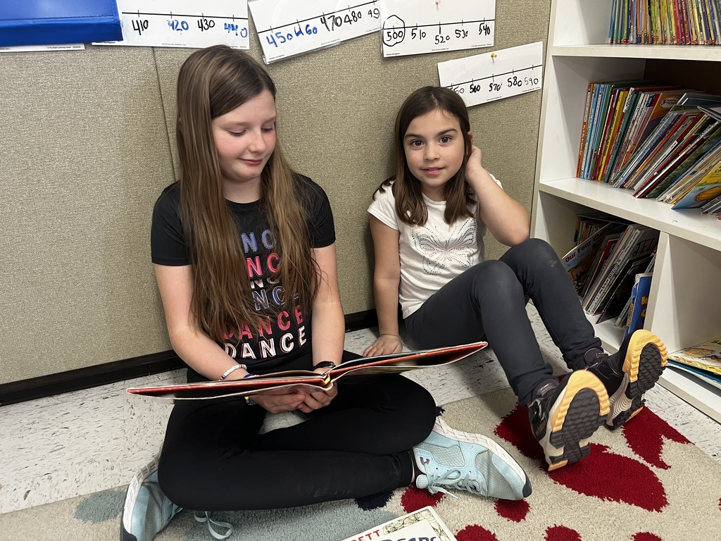 Two young girls are sitting on a patterned rug on the classroom floor, leaning against a beige wall. The girl on the left, wearing a black "DANCE" t-shirt, holds a wide picture book open across her lap as she reads. The girl on the right, in a white butterfly shirt, looks toward the camera. A bookshelf filled with colorful children's books is visible to their right, and hand-written number lines are pinned to the wall above them.