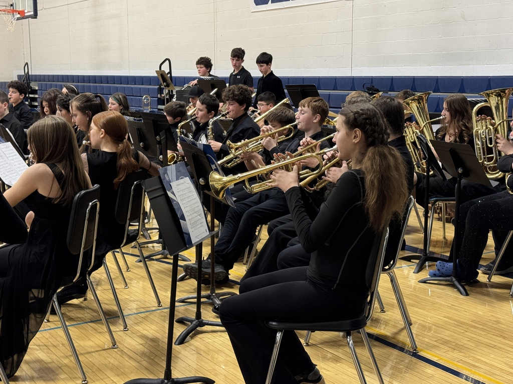 Middle school and high school band students play trumpets and saxophones, focused on their sheet music during the performance.