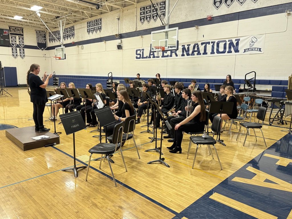 Wide shot of the full band seated across the gym floor as the conductor leads from a podium under a “Slater Nation” banner.