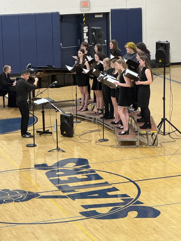Students in the choir sing from risers while the conductor gestures with raised hands; audience members watch from nearby seats.