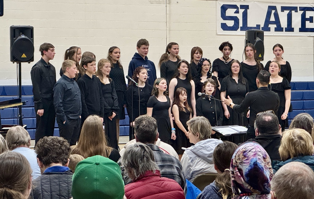 Middle and high school choir dressed in black performing on risers while a conductor leads them from the front of the gym.