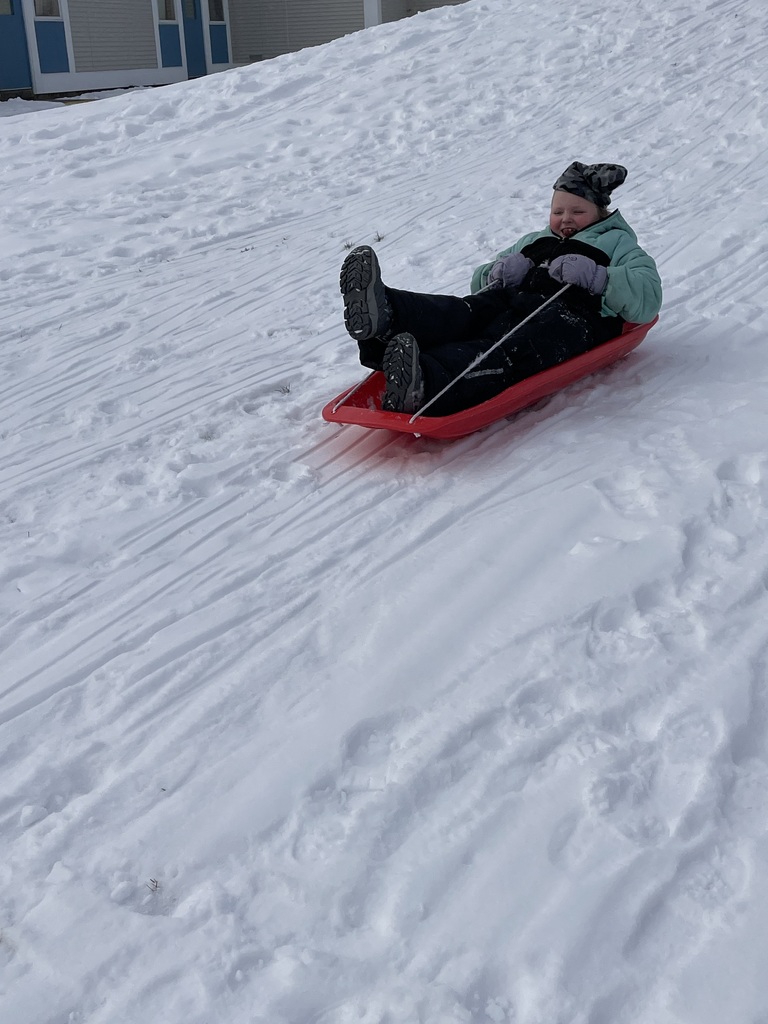 A child wearing a light green jacket, black snow pants, and a camouflage beanie sits in a red plastic sled. They are sliding down a snowy hill, holding onto the rope handles with purple gloves and smiling.