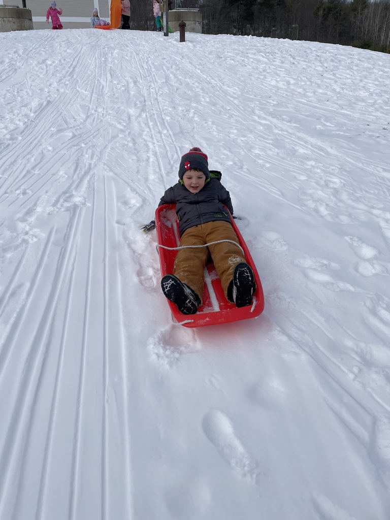 A young boy wearing a grey jacket, tan pants, and a Spiderman-themed beanie sits in a red sled. He is looking down at the snow as he slides, with deep sled tracks visible on the hill behind him.