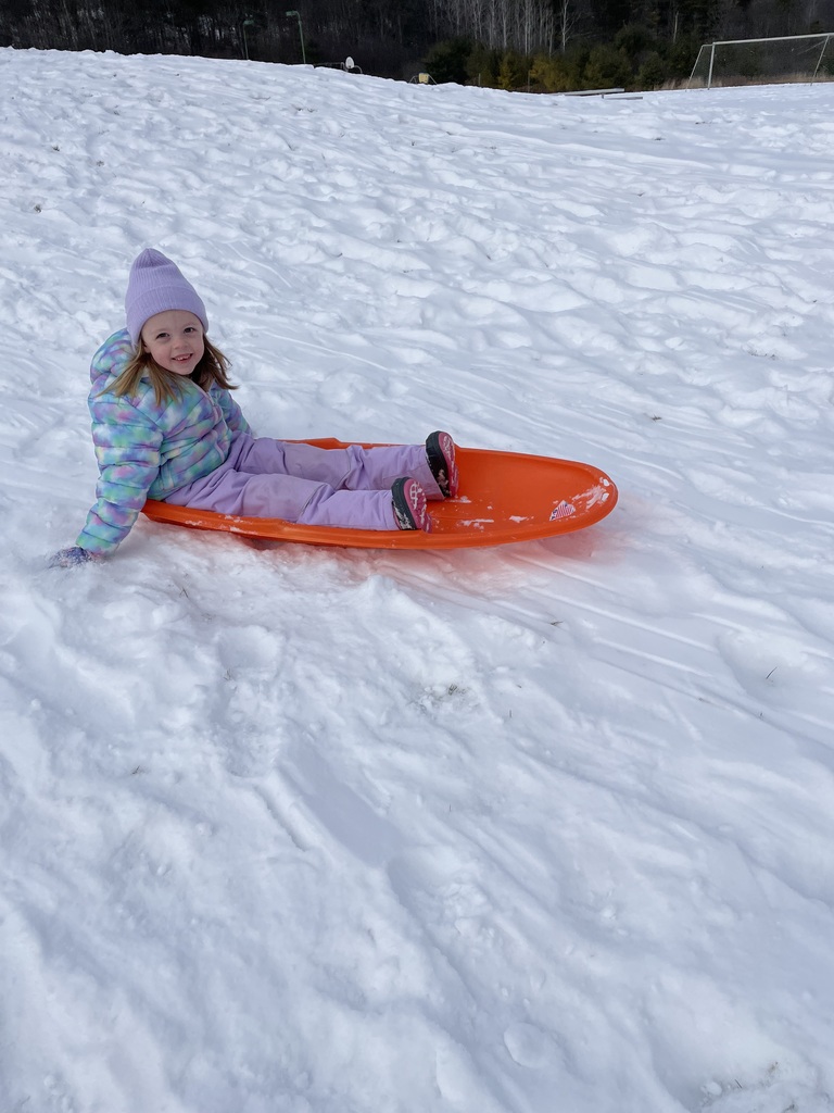 A young child in a blue puffy jacket and a camouflage hat waves one hand while sliding down a snowy slope in a blue sled. A light-colored building with blue trim is visible in the background.