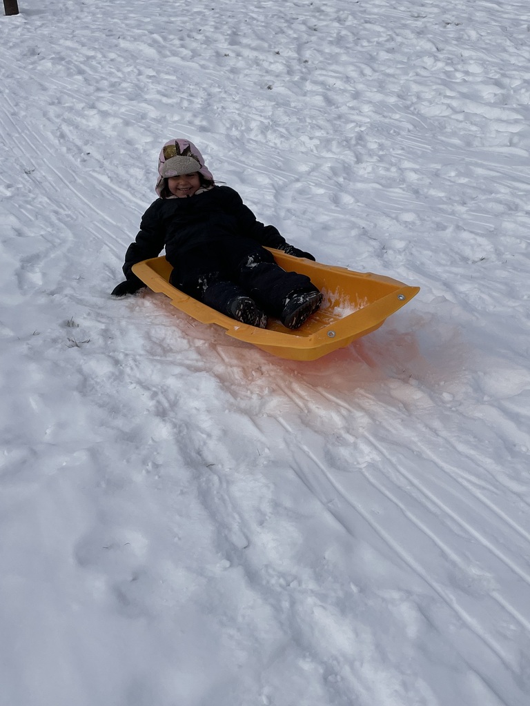 A young girl in a pink coat and a white beanie with a pink bow sits in an orange sled, holding the rope and laughing as she descends a snowy hill. Other children on sleds are visible at the top of the hill in the background.