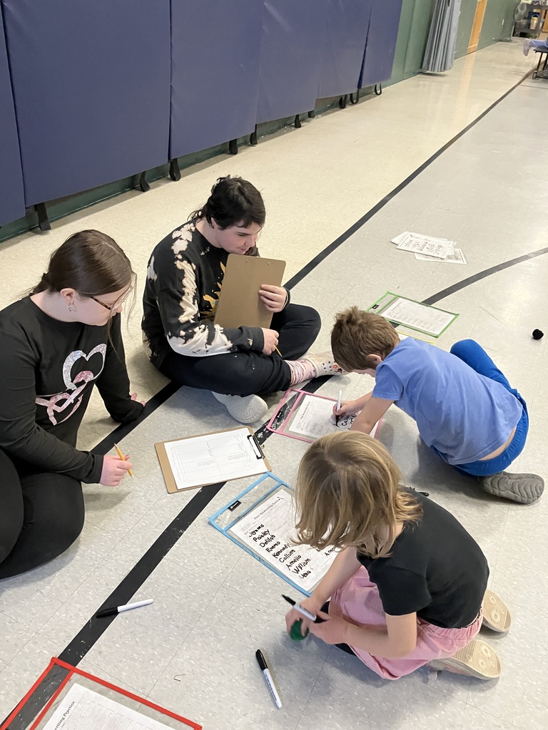 Two older students and two younger children sit in a circle on a gymnasium floor. They are working together on "Fast Word Writing" worksheets placed in plastic sleeves, using dry-erase markers and clipboards to practice writing names and words.