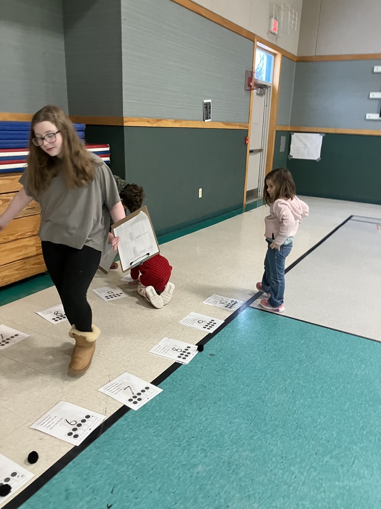 In a gym, an older student walks past a line of numbered papers taped to the floor. Each paper shows a number and a corresponding ten-frame diagram. A younger child in a red dress kneels to interact with one of the papers while another child stands nearby.