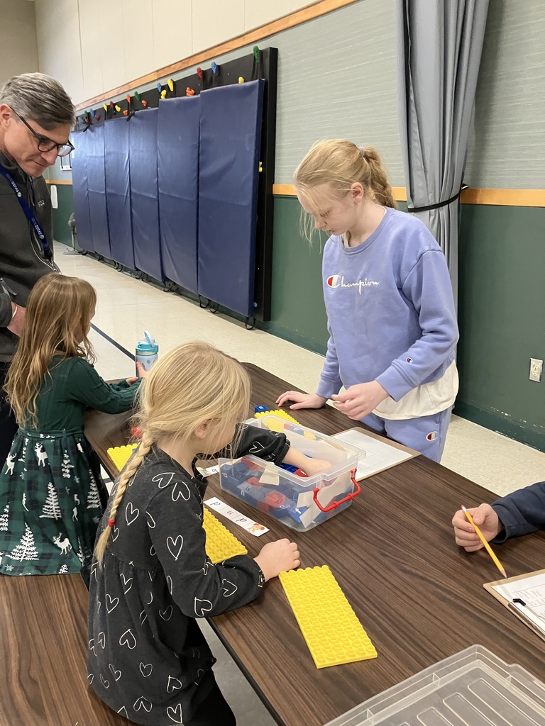 A teenage girl in a light blue sweatshirt leans over a cafeteria table, pointing to lettered blocks to help a young girl in a green dress. They are using yellow baseplates and blue/red blocks to build words. An adult man watches the activity from the side.