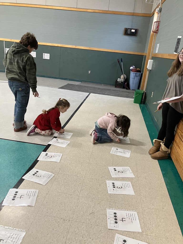 n a gym, an older student walks past a line of numbered papers taped to the floor. Each paper shows a number and a corresponding ten-frame diagram. A younger child in a red dress kneels to interact with one of the papers while another child stands nearby. IMG_2943.jpg A teenage girl in a light blue sweatshirt leans over a cafeteria table, pointing to lettered blocks to help a young girl in a green dress. They are using yellow baseplates and blue/red blocks to build words. An adult man watches the activity from the side.