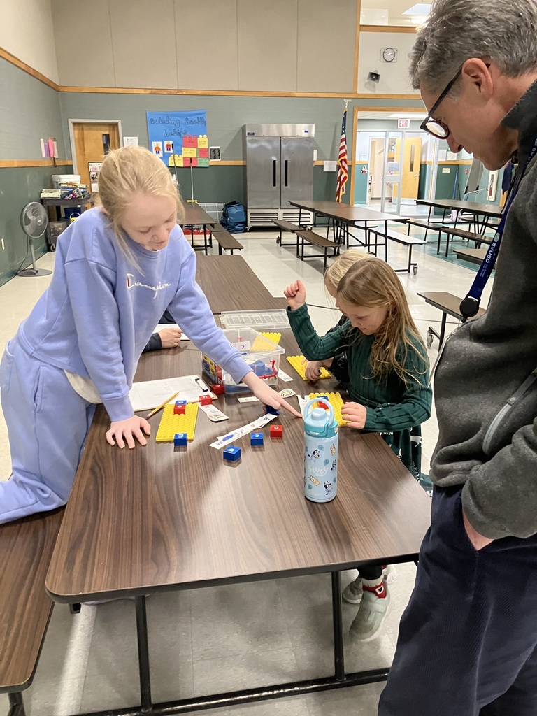 A teenage girl in a light blue sweatshirt leans over a cafeteria table, pointing to lettered blocks to help a young girl in a green dress. They are using yellow baseplates and blue/red blocks to build words. An adult man watches the activity from the side. IMG_2944.jpg A side view of the same literacy activity at the cafeteria table. A young girl with a long blonde braid reaches into a clear bin of letter blocks while a teenage mentor stands by to assist. Yellow building plates and word slips are spread across the table.