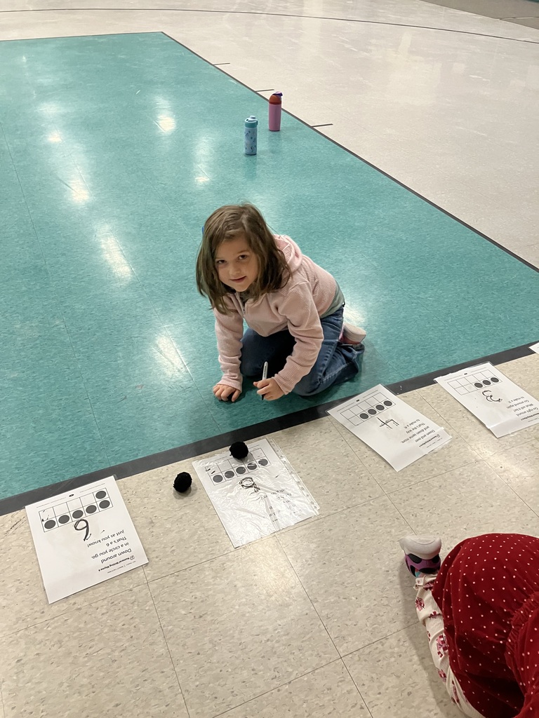 A young girl in a pink hoodie kneels on a green gym floor mat. She is using a dry-erase marker to practice writing the number 6 on a worksheet that includes a ten-frame and a "number rhyme" poem.