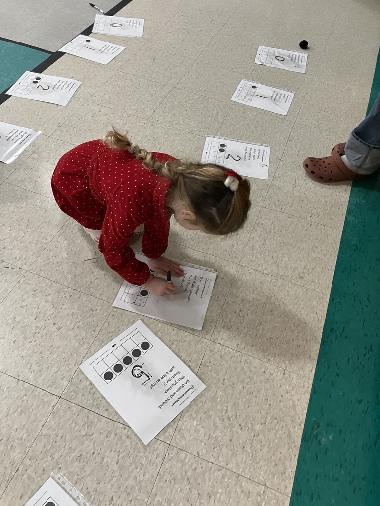 A young girl with a long blonde braid, wearing a red polka-dot dress, kneels on the floor to practice writing the number 5 on a math worksheet. Other numbered stations are visible nearby.