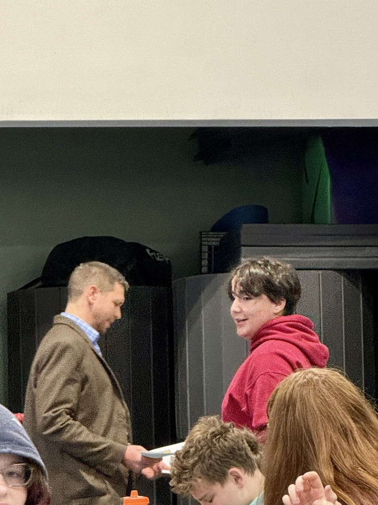 A staff member presents a paper to a student in a red sweatshirt while classmates sit nearby watching.