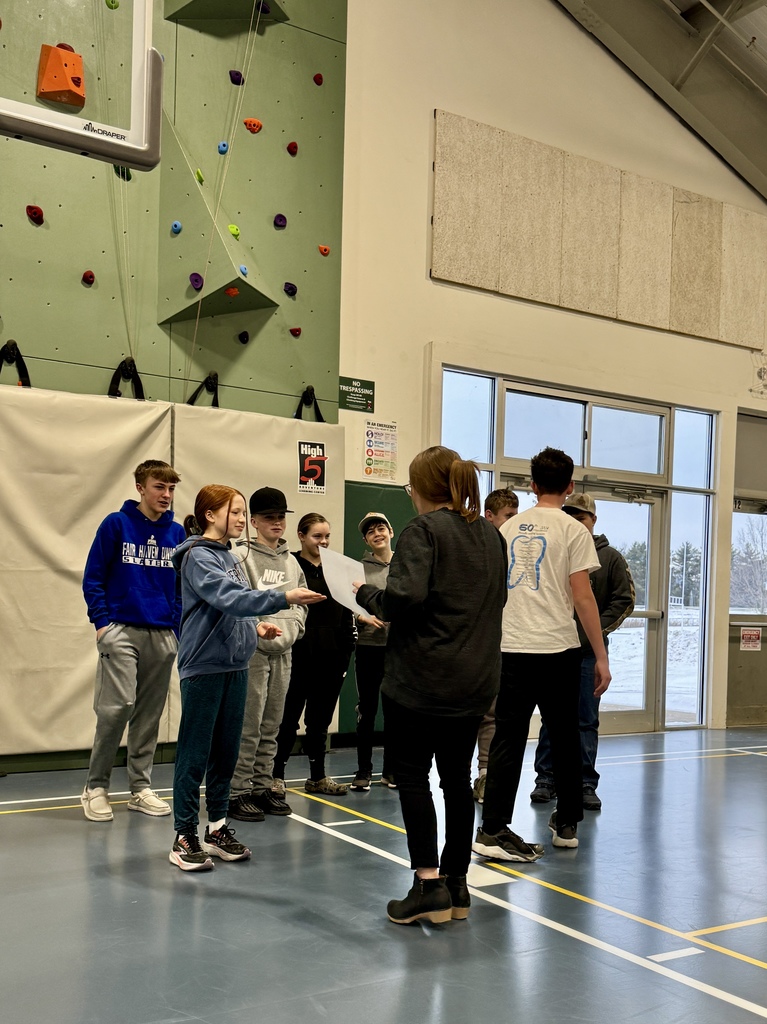 A staff member hands a paper to a student while several students stand in a line waiting their turn during the assembly.