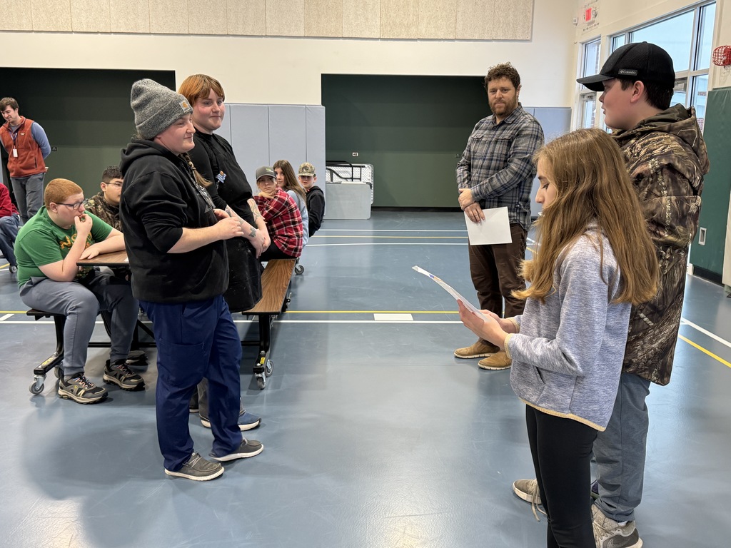 A student reads from a paper while standing in front of peers and staff during an assembly in the gym.