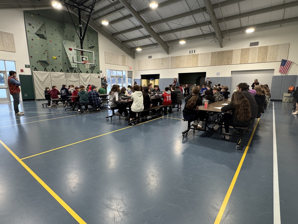 Middle school students sit at cafeteria tables in the gym during an assembly, with staff standing at the front addressing the group.