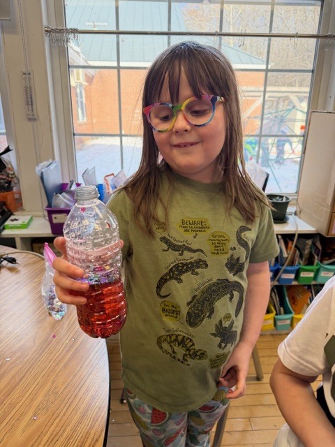 Student wearing glasses smiles while holding up a clear plastic bottle filled with red liquid and colorful materials inside.