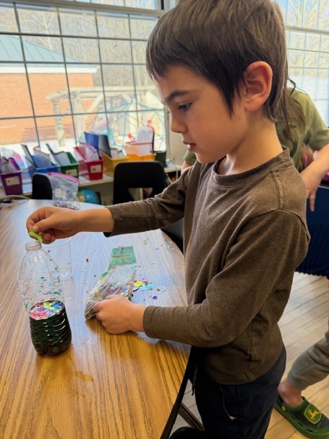 Student adds small materials into a clear plastic bottle on a classroom table, focused on a science experiment with liquids and solids.