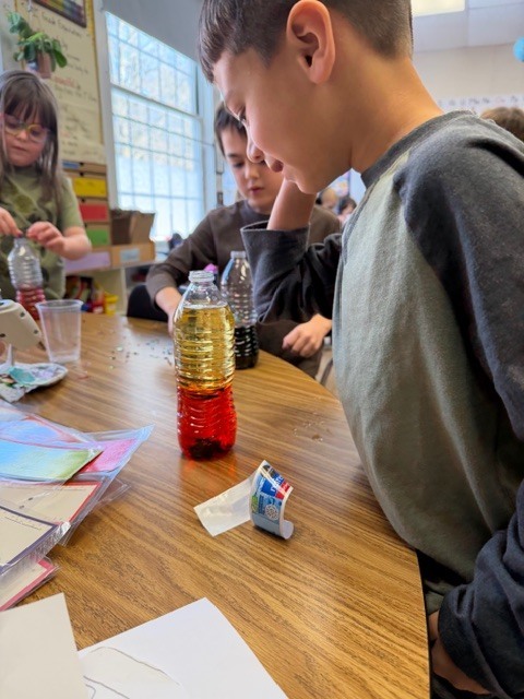 Student leans over a table observing a clear plastic bottle filled with layered red and yellow liquid during a classroom experiment.