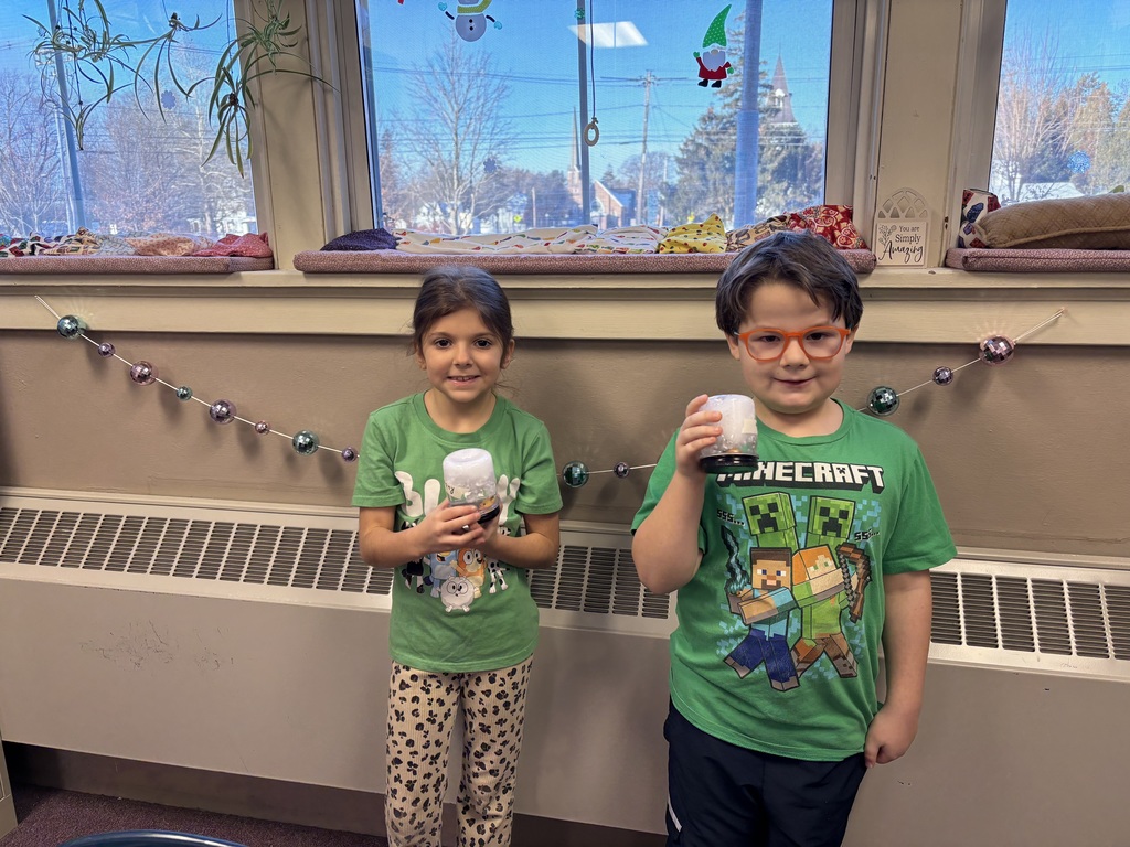 Two students stand side by side holding finished winter-themed craft jars, smiling proudly in front of a classroom window.
